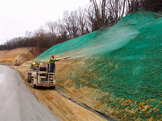 A worker on a truck spraying a green mixture of hydro-mulch onto a burnt hillside. 