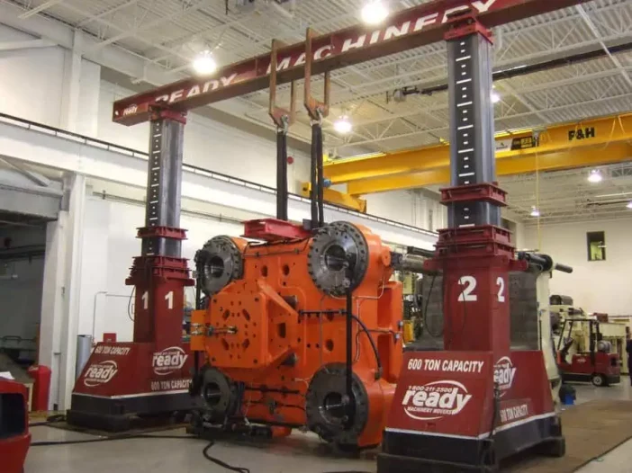 A large industrial machine is positioned in a spacious workshop. The machine features a bright orange central component with multiple circular parts and connections. It is supported by two robust vertical columns labeled "1" and "2," which are painted red and indicate a lifting capacity of 600 tons. Above the machine, a horizontal beam connects the two columns, and there are cables and pulleys attached for lifting purposes. The workshop has high ceilings and is well-lit, with additional machinery visible in the background.