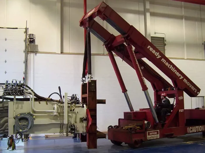 A large red machinery mover is positioned in a spacious industrial setting. The equipment features an articulated arm with a lifting mechanism, currently hoisting a heavy object secured with straps. The machinery mover is equipped with various hydraulic lines and controls, indicating its operational capabilities. A person in a hard hat is seated in the operator's area, overseeing the lifting process. The background includes a white wall and overhead lighting, typical of a warehouse or workshop environment.